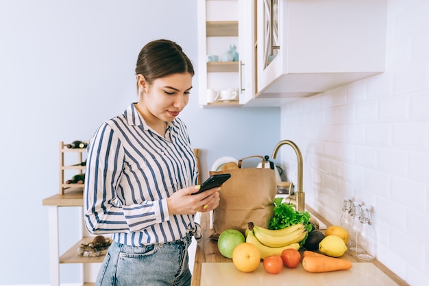 Jovem mulher caucasiana sorridente usar smartphone na cozinha moderna, bolsa com vegetais frescos na mesa. Compra online de alimentos e mantimentos.