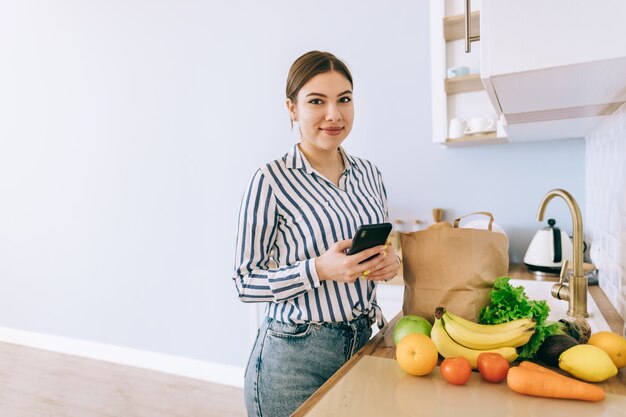 Jovem mulher caucasiana sorridente usar smartphone na cozinha moderna, bolsa com vegetais frescos na mesa. Compra online de alimentos e mantimentos.