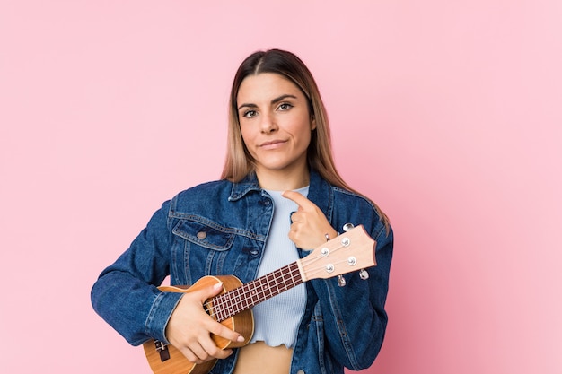 Jovem mulher caucasiana, tocando ukelele apontando com o dedo para você, como se convidando se aproximar.