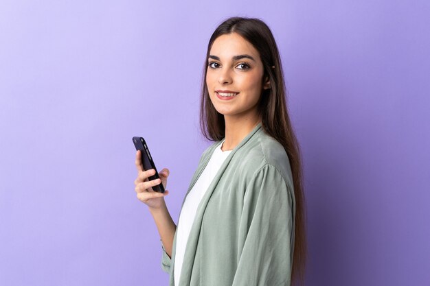 Jovem mulher caucasiana usando celular isolada em roxo sorrindo muito