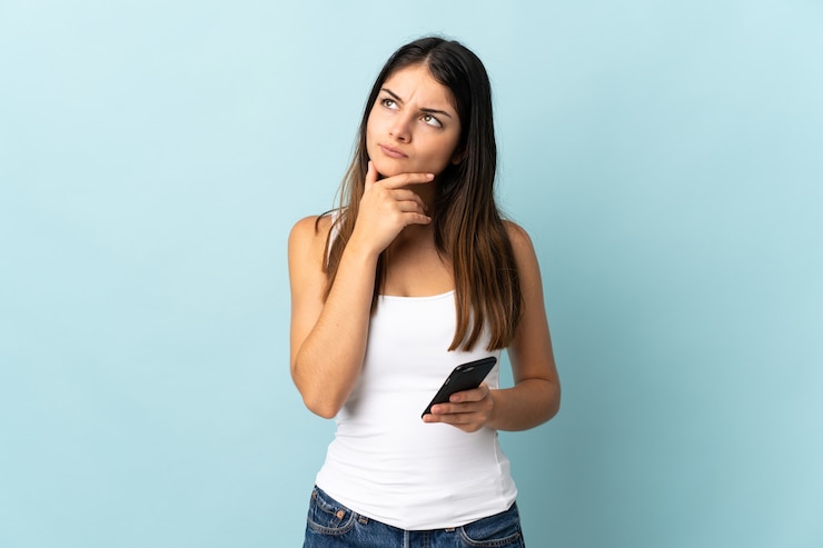 Foto jovem mulher caucasiana usando celular isolado em fundo azul, tendo dúvidas