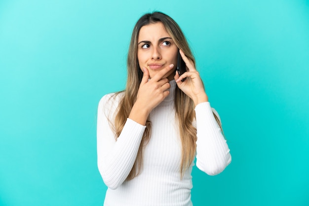 Jovem mulher caucasiana usando celular isolado em fundo azul, tendo dúvidas