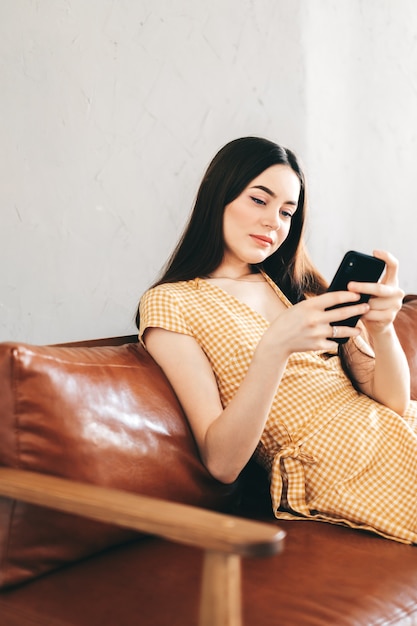 Jovem mulher caucasiana usando smartphone enquanto descansava em um elegante sofá em casa.