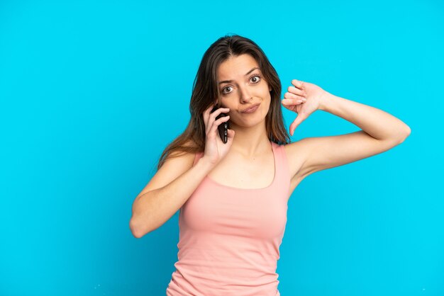 Jovem mulher caucasiana usando telefone celular isolado em fundo azul, mostrando o polegar para baixo com expressão negativa