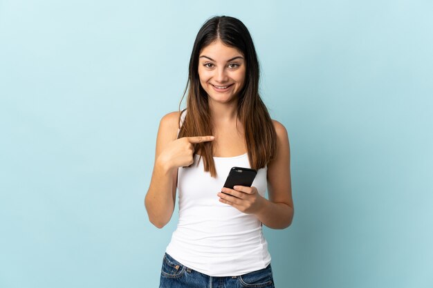 Jovem mulher caucasiana usando telefone celular isolado em um fundo azul com expressão facial surpresa