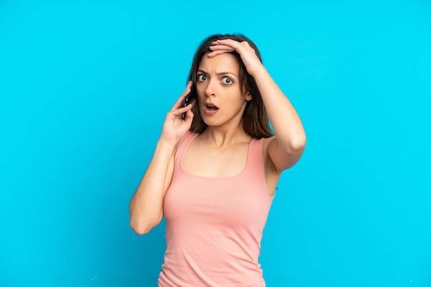 Jovem mulher caucasiana usando telefone celular isolado em um fundo azul, fazendo gesto surpresa enquanto olha para o lado