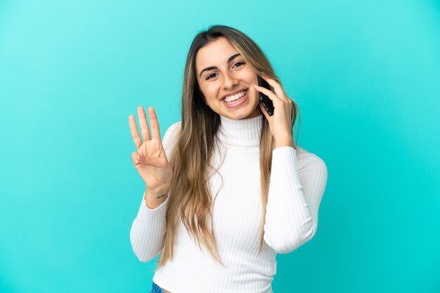 Jovem mulher caucasiana usando telefone celular isolado em um fundo azul feliz e contando três com os dedos