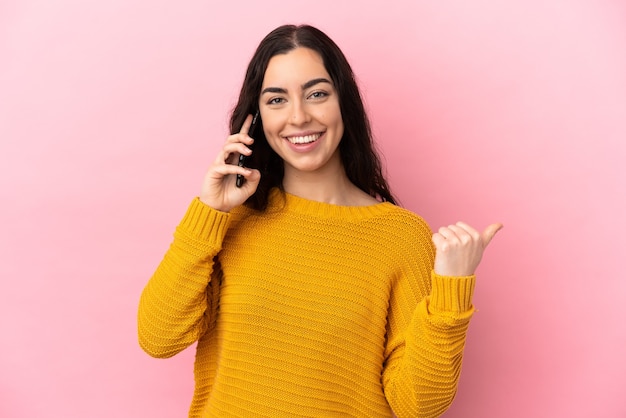 Jovem mulher caucasiana usando telefone celular isolado em um fundo rosa apontando para o lado para apresentar um produto