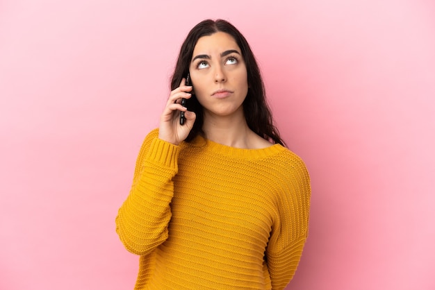 Jovem mulher caucasiana usando telefone celular isolado em um fundo rosa e olhando para cima