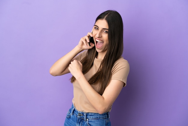 Jovem mulher caucasiana, usando telefone celular isolado em um fundo roxo, comemorando uma vitória