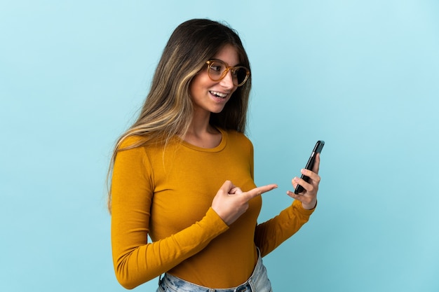 Foto jovem mulher caucasiana usando telefone celular isolado em uma parede azul apontando para o lado para apresentar um produto