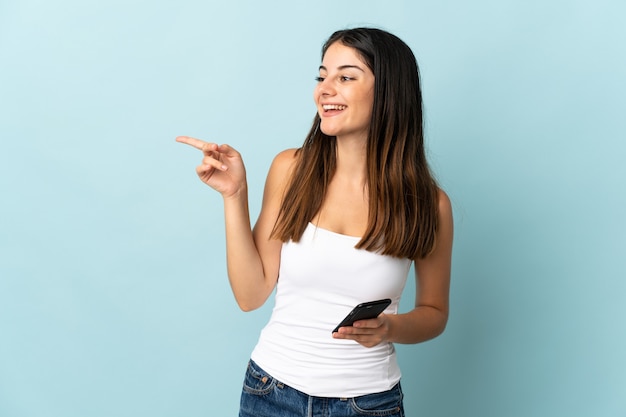 Jovem mulher caucasiana usando telefone celular isolado na parede azul apontando o dedo para o lado e apresentando um produto