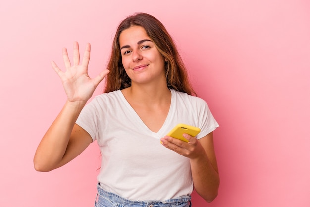 Foto jovem mulher caucasiana, usando telefone celular isolado no fundo rosa, sorrindo alegre mostrando o número cinco com os dedos.