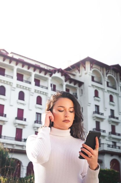 Jovem mulher caucasiana usando um smartphone em frente a um prédio branco