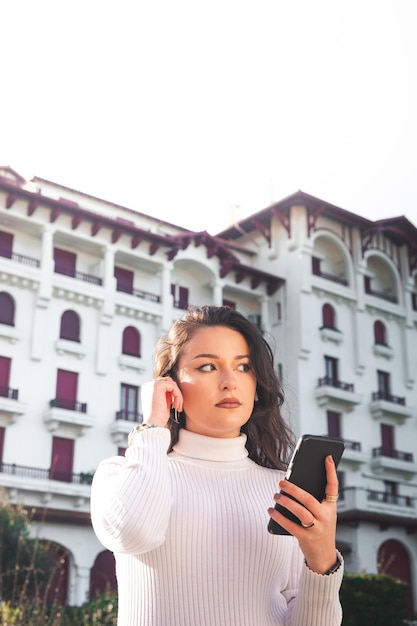 Jovem mulher caucasiana usando um smartphone em frente a um prédio branco