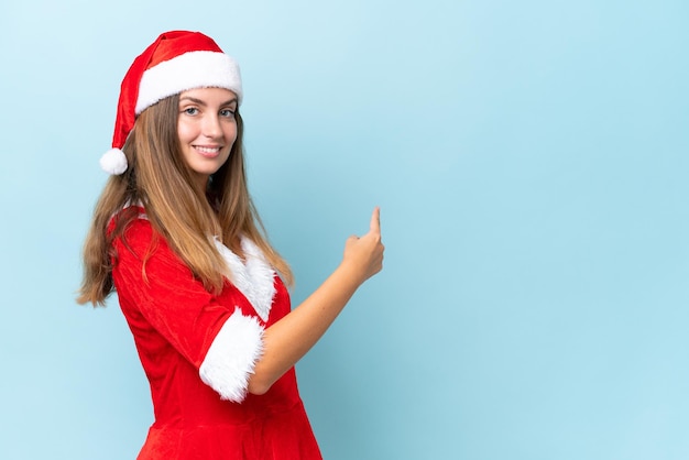 Jovem mulher caucasiana vestida como mamãe noel isolada em fundo azul apontando para trás