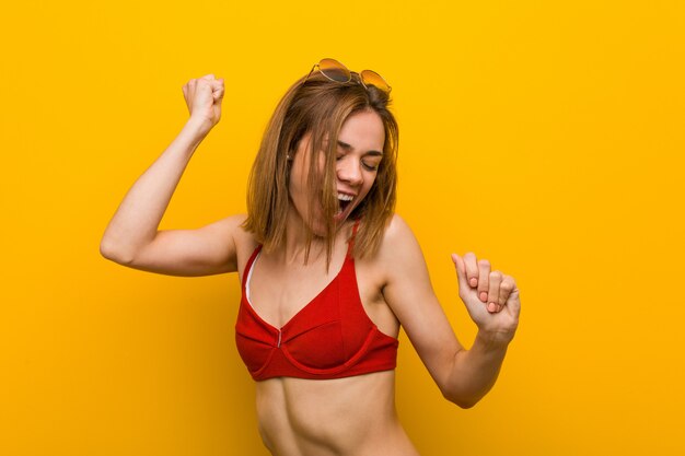 Jovem mulher caucasiana, vestindo biquíni e óculos de sol, comemorando um dia especial, saltos e levante os braços com energia.