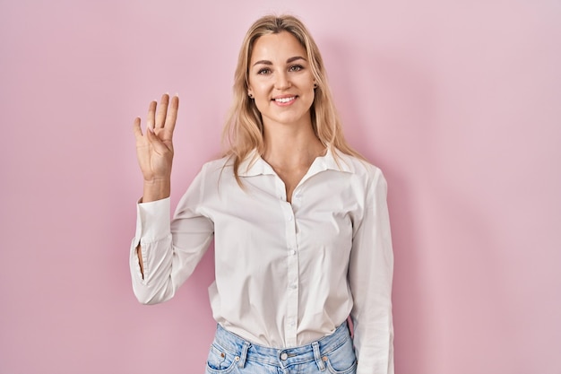 Jovem mulher caucasiana vestindo camisa branca casual sobre fundo rosa mostrando e apontando para cima com os dedos número quatro enquanto sorria confiante e feliz.