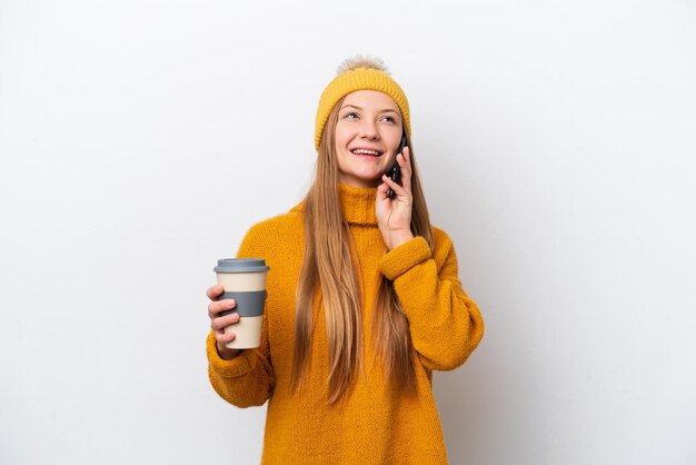 Jovem mulher caucasiana vestindo jaqueta de inverno isolada em fundo branco segurando café para levar e um celular