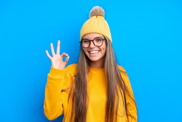 Jovem mulher caucasiana vestindo roupas de inverno isoladas em fundo azul, mostrando sinal de ok com os dedos