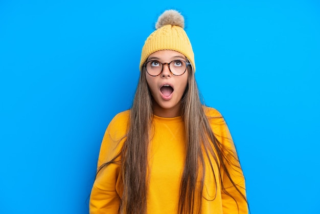 Jovem mulher caucasiana vestindo roupas de inverno isoladas em fundo azul, olhando para cima e com expressão de surpresa