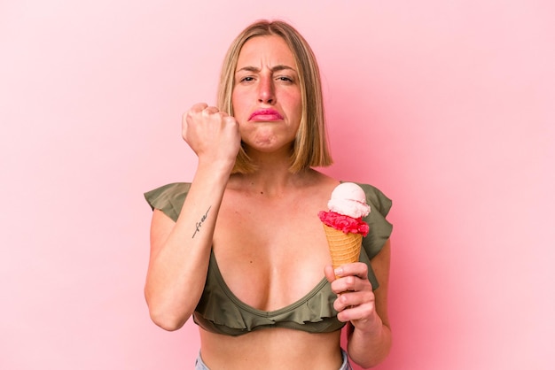 Jovem mulher caucasiana, vestindo um biquíni e segurando um sorvete isolado no fundo rosa, mostrando o punho para a expressão facial agressiva da câmera