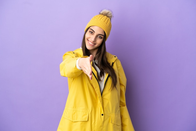 Jovem mulher caucasiana vestindo um casaco à prova de chuva isolado em um fundo roxo apertando as mãos para fechar um bom negócio