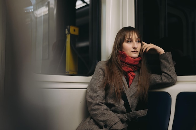 Jovem mulher caucasiana, vestindo um casaco elegante e lenço, sentada sozinha no vagão do metrô