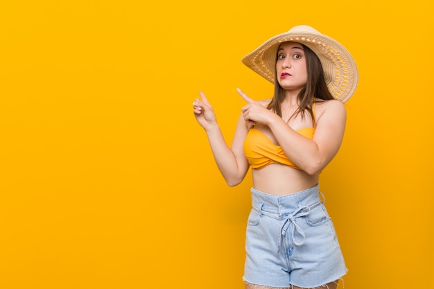 Jovem mulher caucasiana, vestindo um chapéu de palha, olhar de verão chocado apontando com o dedo indicador para um espaço em branco.