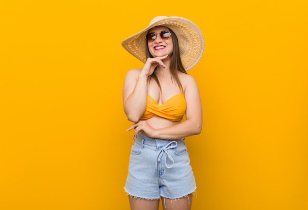Jovem mulher caucasiana, vestindo um chapéu de palha, olhar de verão sorrindo feliz e confiante, tocando o queixo com a mão.