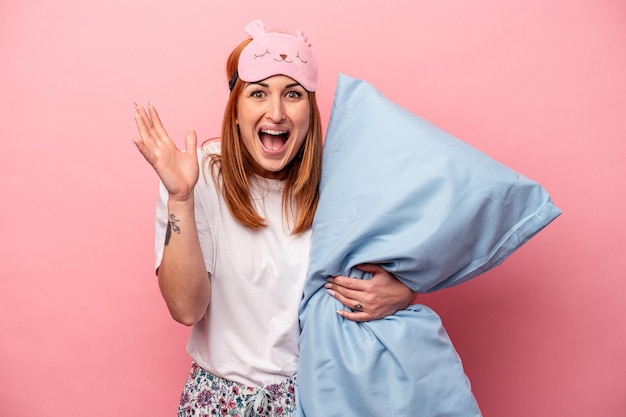 Jovem mulher caucasiana, vestindo um pijama segurando o travesseiro isolado no fundo rosa, recebendo uma surpresa agradável, animado e levantando as mãos.