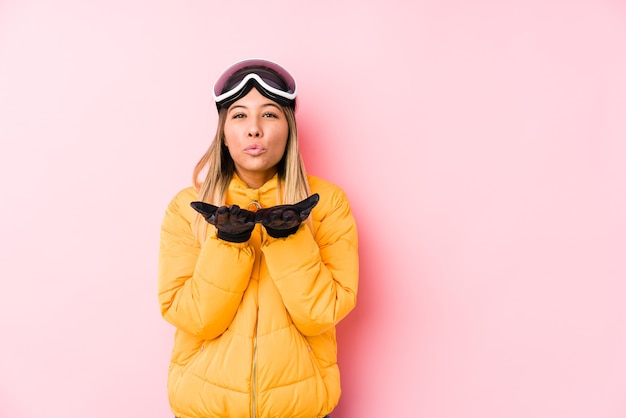 Jovem mulher caucasiana, vestindo uma roupa de esqui em uma parede rosa, dobrando os lábios e segurando as palmas das mãos para enviar beijo no ar.