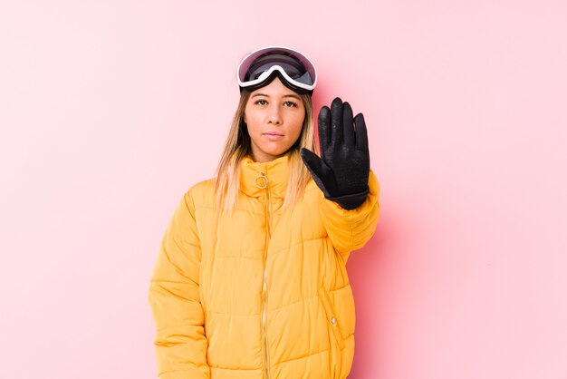 Jovem mulher caucasiana, vestindo uma roupa de esqui em uma parede rosa em pé com a mão estendida, mostrando o sinal de stop, impedindo-o.