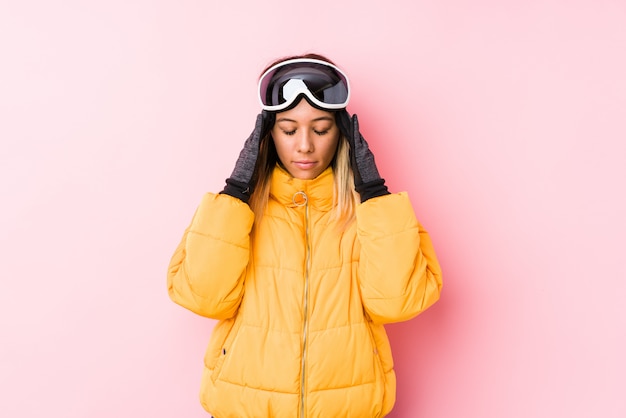 Jovem mulher caucasiana, vestindo uma roupa de esqui em uma parede rosa tocando templos e tendo dor de cabeça.