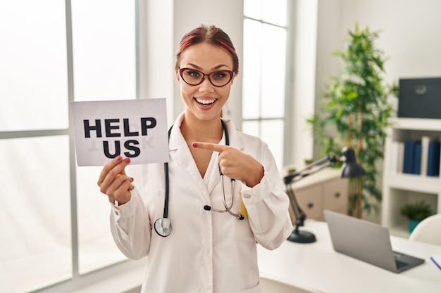 Jovem mulher caucasiana vestindo uniforme médico segurando ajuda nos banner sorrindo feliz apontando com a mão e o dedo