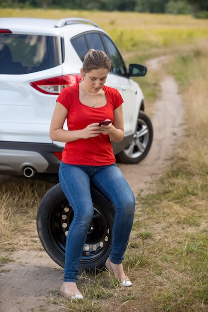 Foto jovem mulher chateada sentando em um pneu sobressalente ao lado de um carro quebrado em campo e procurando por um número de telefone de serviço