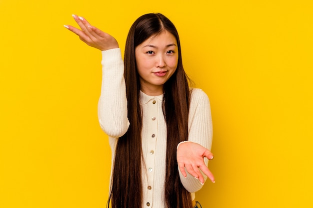 Jovem mulher chinesa isolada em fundo amarelo faz escala com os braços, sente-se feliz e confiante.