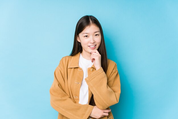 Jovem mulher chinesa posando em azul isolada sorrindo feliz e confiante, tocando o queixo com a mão.