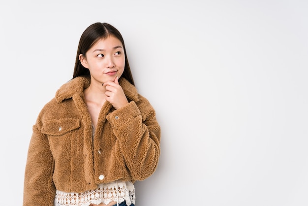 Jovem mulher chinesa posando em um branco isolado, olhando de soslaio com expressão duvidosa e cética.