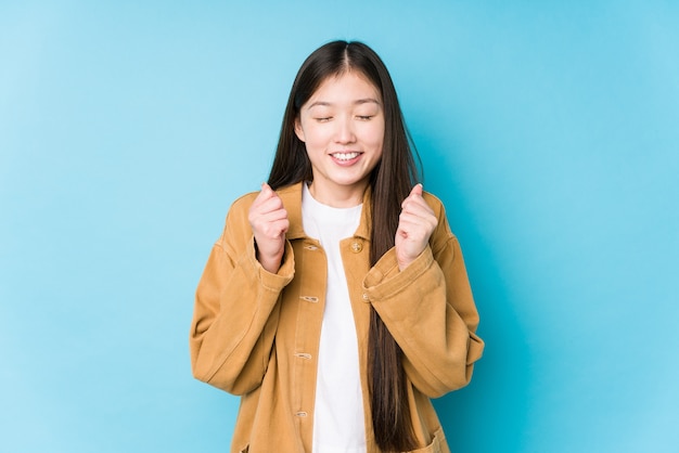 Jovem mulher chinesa posando em um fundo azul isolado levantando o punho, sentindo-se feliz e bem sucedido. Conceito de vitória.