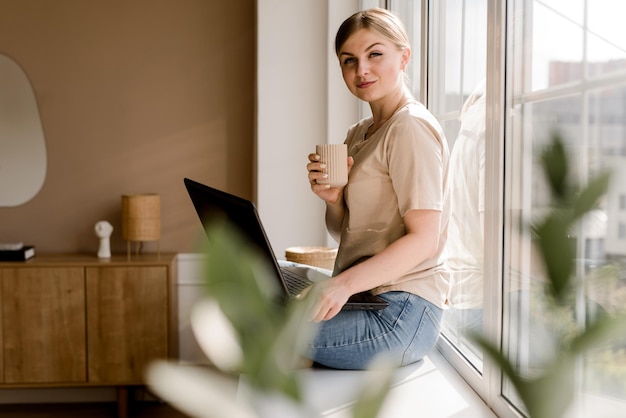 Jovem mulher com a xícara de café e seu laptop sentado no parapeito da janela