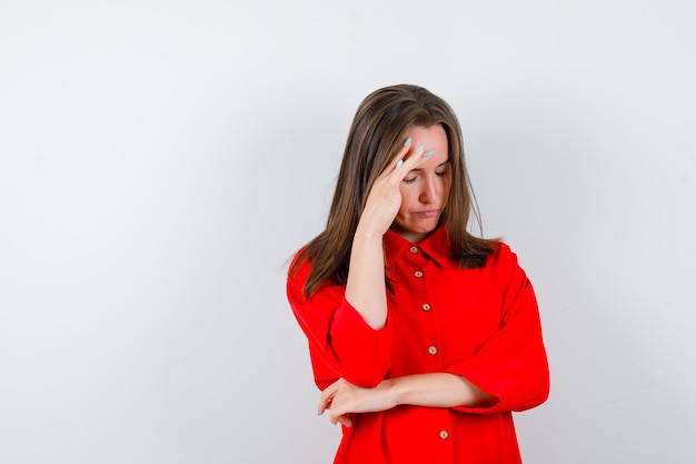 Foto jovem mulher com as mãos no rosto, fechando os olhos na blusa vermelha e parecendo chateada, vista frontal.