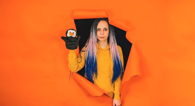 Jovem mulher com brinquedo de fantoche macio na mão olhando para fora do buraco de fundo laranja Muito fêmea com pinguim de fantoche Conceito de show de marionetes