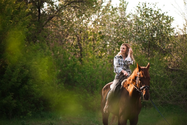 Jovem mulher com cabelo comprido, posando com um cavalo castanho em uma floresta em um prado ensolarado.