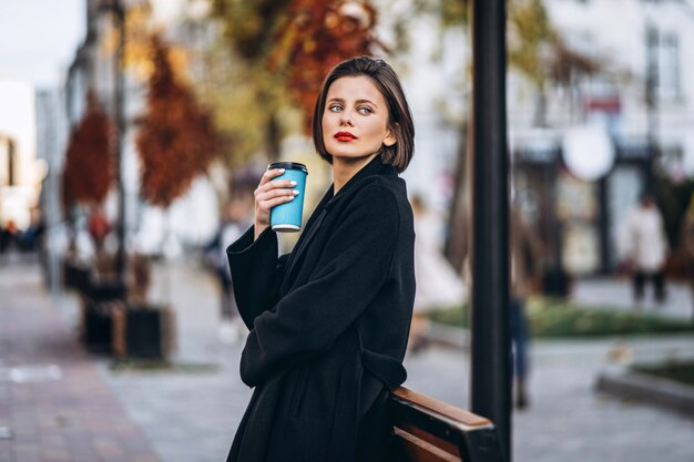 Jovem mulher com cabelo curto e lábios vermelhos, segura uma xícara de café ao ar livre