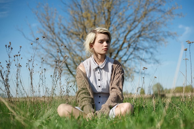 Jovem mulher com cabelo curto em um vestido manchado e suéter sentada no chão em campo em dia de sol