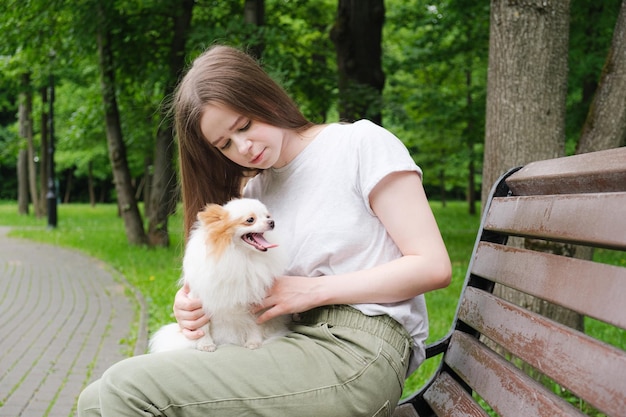 Jovem mulher com cabelo longo e reto se senta em um banco no parque e segura um cachorrinho fofo em seus braços