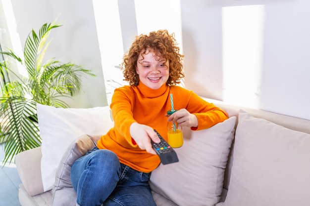 Jovem mulher com cabelo ruivo cacheado, segurando o controle remoto e assistindo tv, mudando de canal. Linda garota bebendo suco de laranja e assistindo tv no apartamento dela