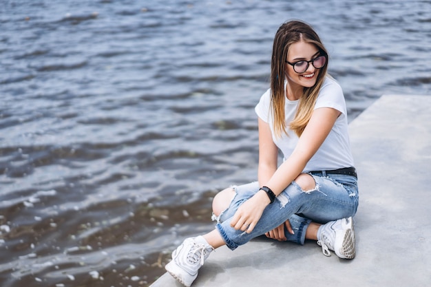 Jovem mulher com cabelos longos em copos elegantes posando na margem concreta perto do lago. Garota vestida de jeans e camiseta, sorrindo e olhando para longe