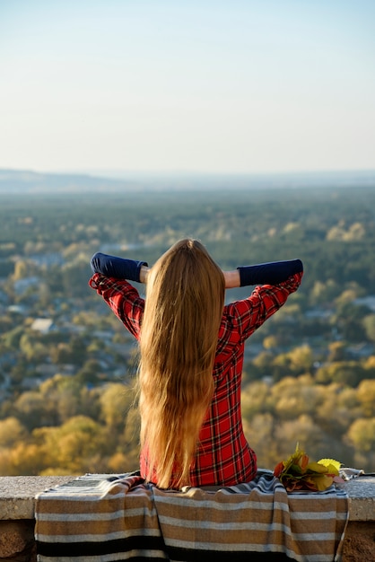 Foto jovem mulher com cabelos longos, senta-se numa colina com vista para a cidade. vista traseira